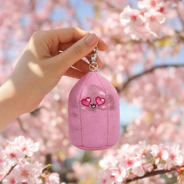 A hand holding Baby Rosie, the pink rose quartz bag charm, in front of cherry blossoms