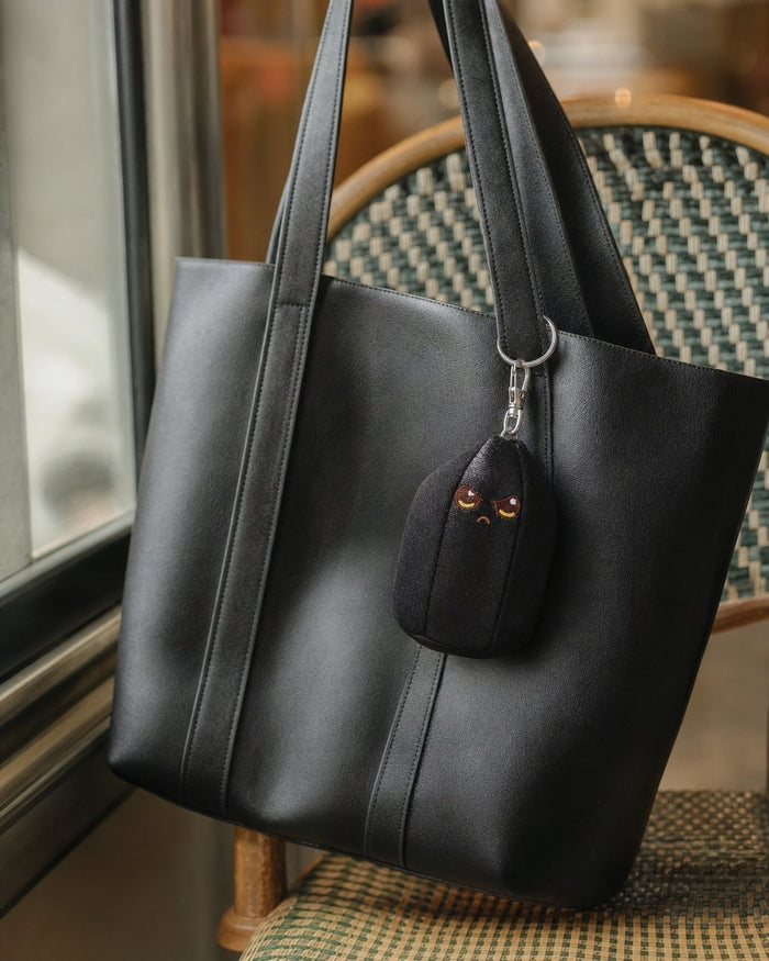 Baby Obie bag charm hanging from a black leather tote on a cafe chair
